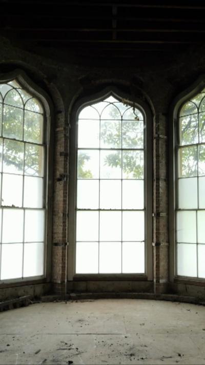 Large and tall windows are pictured in an historic mansion, with bright indirect light beaming through. The floor is dirty and dusty.