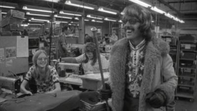 DJ Del Perry stands in a clothing factory wearing a fur-lined coat and holding a cane.