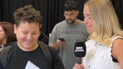 Student in a black tshirt smiles and looks at results paper. He stands next to BBC presenter