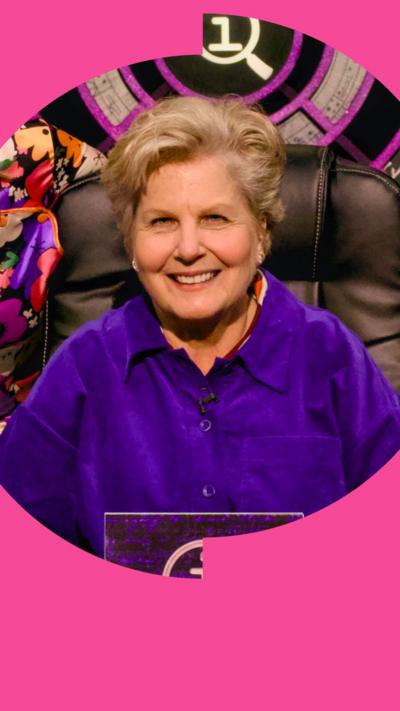 Sandi Toksvig smiles in a purple shirt while sat in a black chair in front of the QI logo 