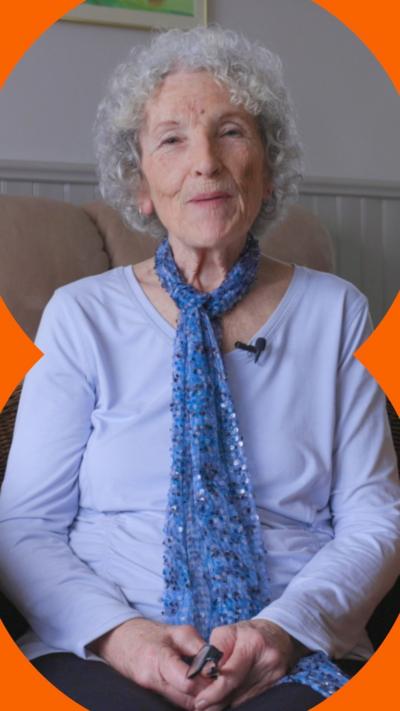 An older woman with curly grey hair, sits in an armchair looking at the camera. She's wearing a lilac top and a blue scarf.