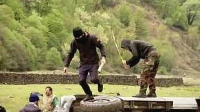 Two hooded men stand atop an overturned car