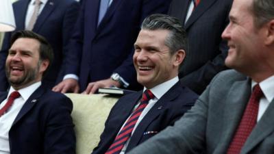 JD Vance, Pete Hegseth and Mike Waltz sit on a yellow sofa wearing suits laughing.