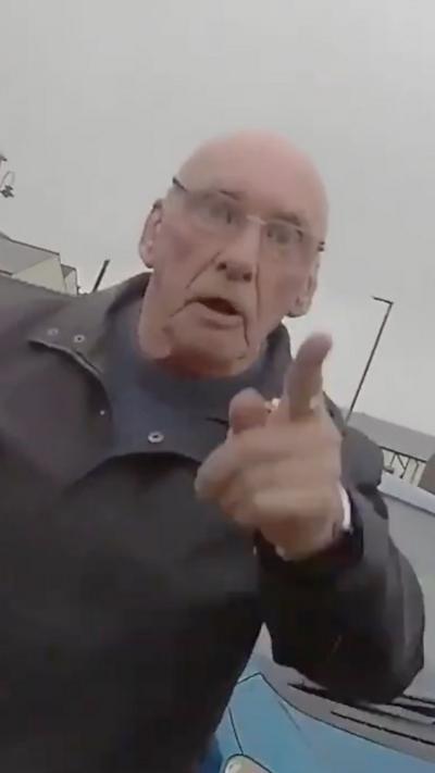 A bald man in glasses pointing in front of a blue car