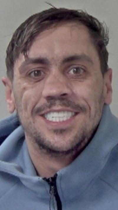 A man in a blue tracksuit with white teeth in a police mugshot.