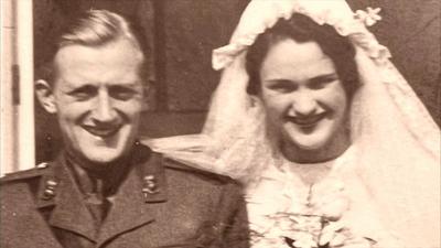A sepia-colour archive photo of Margaret and Rex stood together on some steps outside a building on their wedding day. She is wearing her wedding dress and holding flowers, while he is smiling in military uniform.