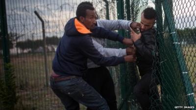 Migrants help a man squeeze through a gap in a fence near the Eurotunnel terminal in Calais