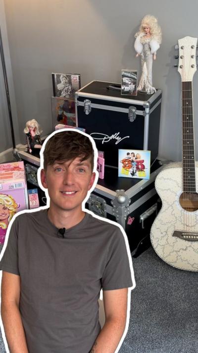 A man with brown hair wearing a grey shirt outlined in white in front of Dolly Parton memorabilia