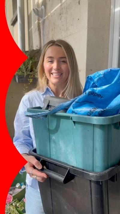 A woman with long blonde hair talks to the camera while holding two recycling tubs