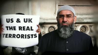 Anjem Choudary with a protestor holding banner stating 'US & UK The Real Terrorists'.