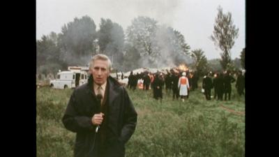 The reporter standing in front of a crowd surrounding the burning plane.