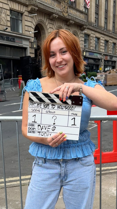 A woman stands in front of a street in the city centre of Glasgow that has been dressed up for a movie set. She has short ginger hair, is wearing a blue top and is holding a wooden director's movie sign.