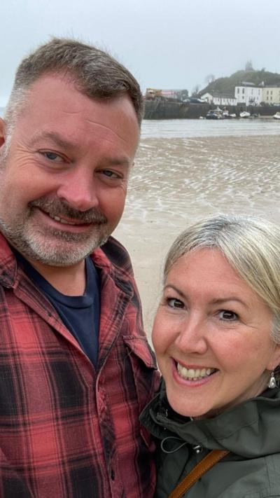 A grey man and a blonde woman stood on a beach with sand and waves behind them.