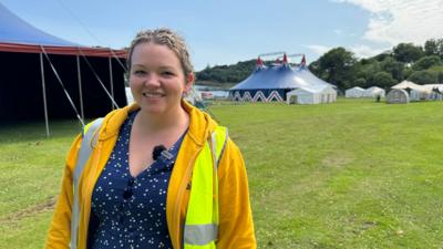 Eilidh Linc, Iar-chathraiche Fèis Cheilteach Innse Gall, na seasamh air làrach na Fèise air là grianach le feur uaine timcheall oirra agus gàire mhòr air a h-aodann.