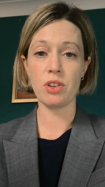 A woman with a short blonde bob and blue eyes wears a black top and grey blazer. She sits in front of a dark green wall, a bookshelf and a picture on the wall.