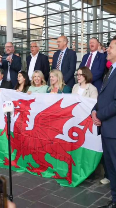 A group of politicians stand on the steps of the Senedd in two rows. The bottom row hold a large Welsh flag. 
