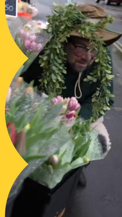 Phillip Norton wearing a hat with green foliage dangling down. He is crouching behind bouquets of flowers.