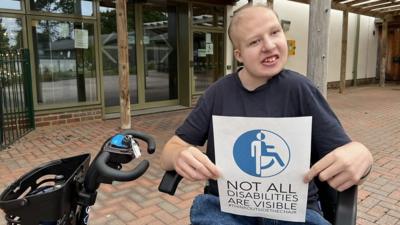 Sam is at in a wheelchair holding a sign that says "not all disabilities are visible"