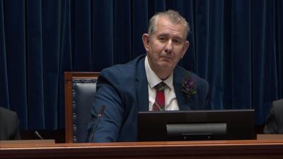 A man sits behind a wooden podium. The top of a computer monitor can be seen over the wooden edge of the podium. The man is sitting on a wooden seat with blue leather. He is wearing a blue suit with a tartan tie. His balding grey hair is slicked back. 