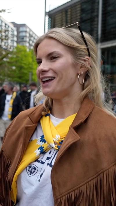 A woman with blonde hair and a brown jacket stands in a white t-shirt and a yellow scarf