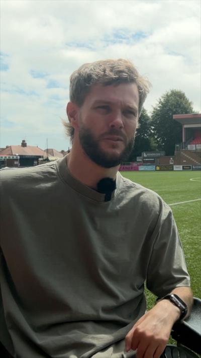 young man with brown hair and beard and green top sat in wheelchair on edge of football pitch