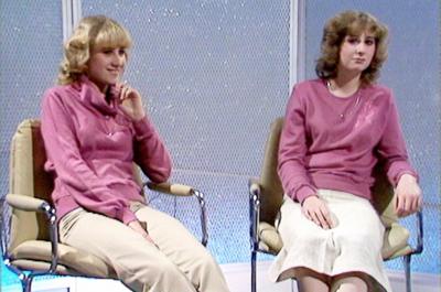 Both twins sitting in the studio. They are both wearing pink jumpers, one with cream trousers and the other with a white skirt.