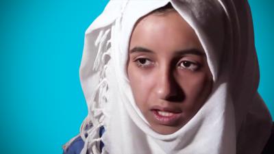 A girl wearing a white headscarf speaks seriously against a blue background