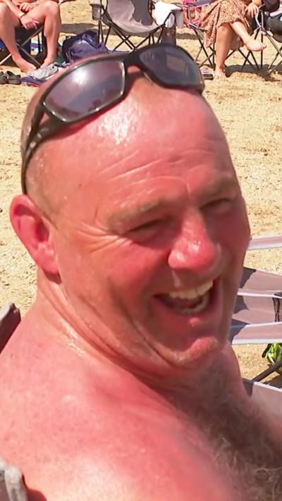 A man is sat in a camping chair laughing. He is wearing sunglasses on top of his head, in the background is sand from the beach and legs of other beach-goers that are not fully in frame