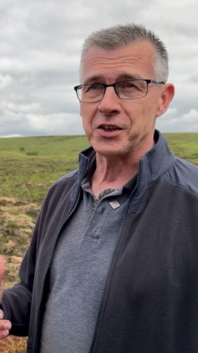 Man with short grey hair and black framed square glasses. He is wearing a navy fleece over a grey polo shirt. Behind him is a grey sky and a mound of grass. 