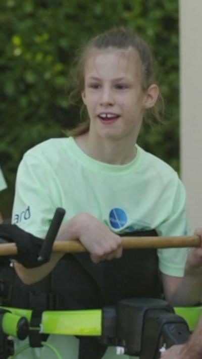 A girl in a T-shirt holding on to a yellow bar that's part of a robotic exoskeleton