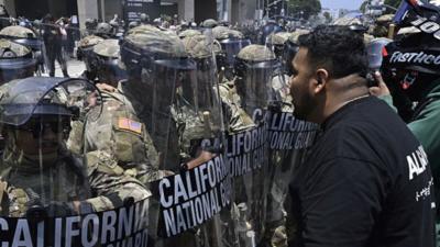 A row of military personnel in camouflage uniforms and helmets stands holding shields as a protester confronts them.