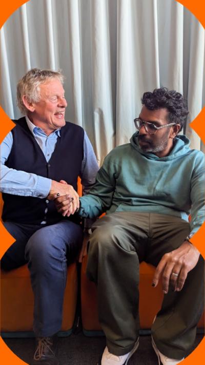 Martin Clunes and Romesh Ranganathan shake hands whilst sat down on a orange sofa.