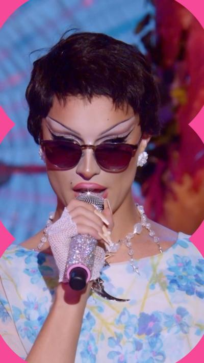 Drag queen Bones wears a floral dress with a short dark wig and sunglasses. She holds a sparkly mic