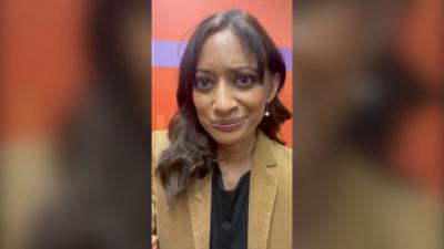 Dharshini David has long brown hair and wears a camel jacket and black shirt