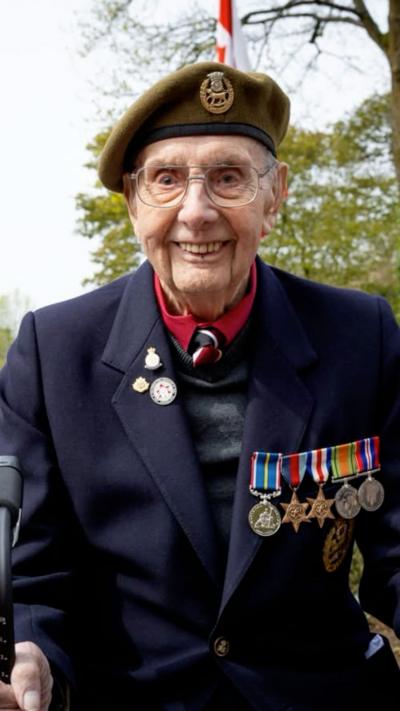 An old man wearing a military beret and jacket with a string of medals smiles for the camera