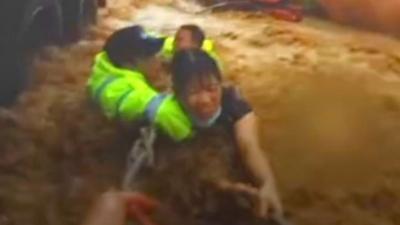 A woman holds onto a rope in rushing flood waters, with rescue workers holding her