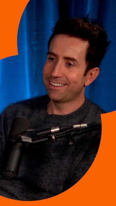 A man, Nick Grimshaw, smiling in a podcast studio, with a blue curtain behind.
