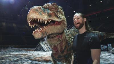 A male puppeteer bears his teeth whilst standing next to a dinosaur on stage