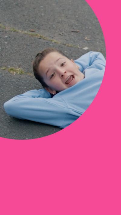A girl in a pale blue jumper lies on the pavement looking cheeky.