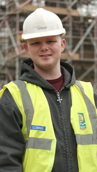 A boy in a white hard hat, a yellow hi-vis, a dark green jacket and maroon t-shirt smiles at the camera. He is standing in front of a building site and scaffolding.