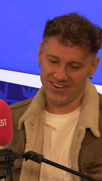 Man with short brown hair speaking in to a pink microphone. He is wearing a grey denim jacket with a fur lining.