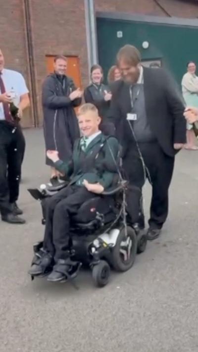 A teenager in a wheelchair, in a playground. Behind him are numerous teachers and friends.