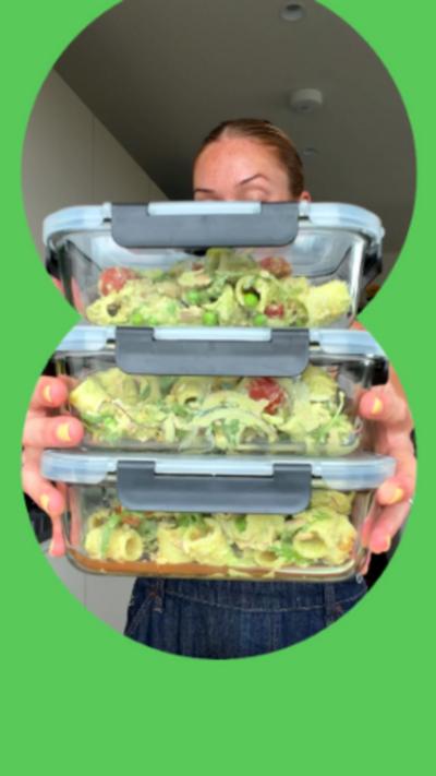 A woman with blonde hair holds three stacked food storage boxes full of a pasta salad with a green sauce.