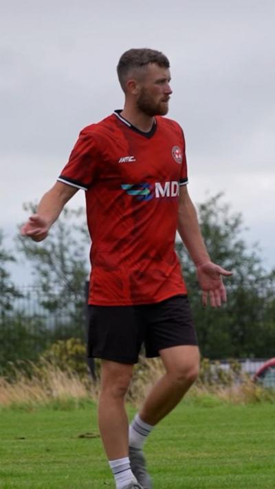 A man wearing an Omagh Town Football Club strip. It consists of a red top with black shorts. He has short brown hair and a short brown beard. He is standing on grass with trees in the background. 