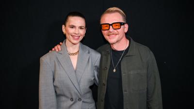 Hayley Atwell in a grey suit and Simon Pegg in a green shirt smile with their arms around each other