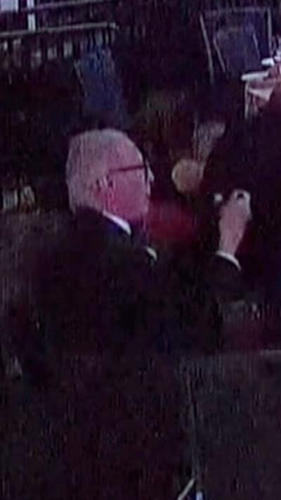 A man with grey hair, wearing a suit, holds a fork up to his mouth, as others are seen crouching or holding up phones