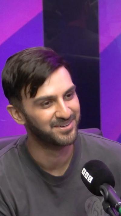 A man with dark hair and facial hair wearing a grey top and smilling