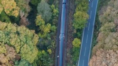 The two trains which collided are visible from above, cutting through autumn trees. To the right is an empty road. 