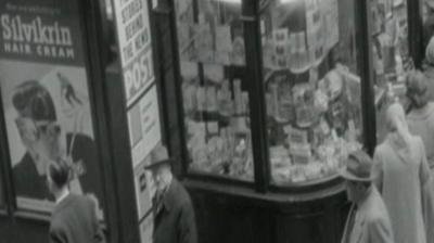 A black and white image of people walking on a street and looking in a shop window