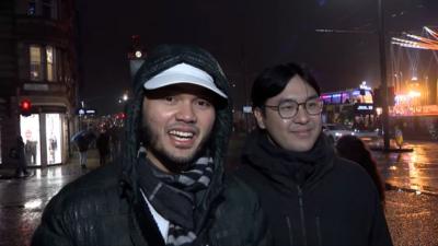 Two men wearing black waterproof jackets stand in the rain on Edinburgh's Princes St as its Festive lights glow in the background. The man on the left wears a white hat under his hood, which is covered in water droplets, and a black and grey scarf. The second man has glasses and a black scarf.